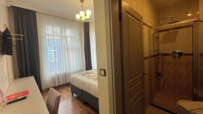 Une chambre d’hôtel équipée d’un lit, d’un bureau, d’une chaise, d’un espace douche et d’une fenêtre avec des rideaux.