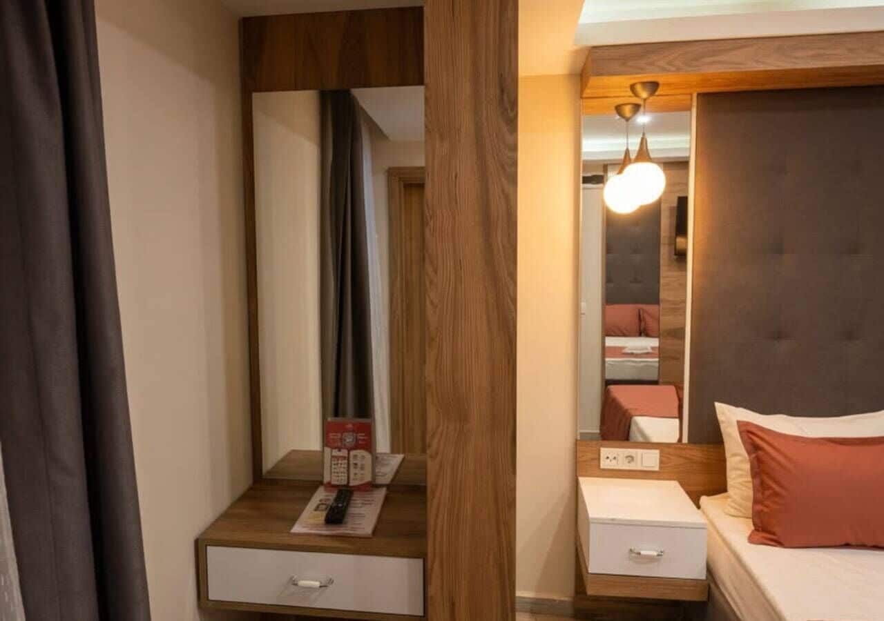 A hotel room with a wooden nightstand, a bedside table, a bed with a headboard, a mirror, and a hanging light fixture.