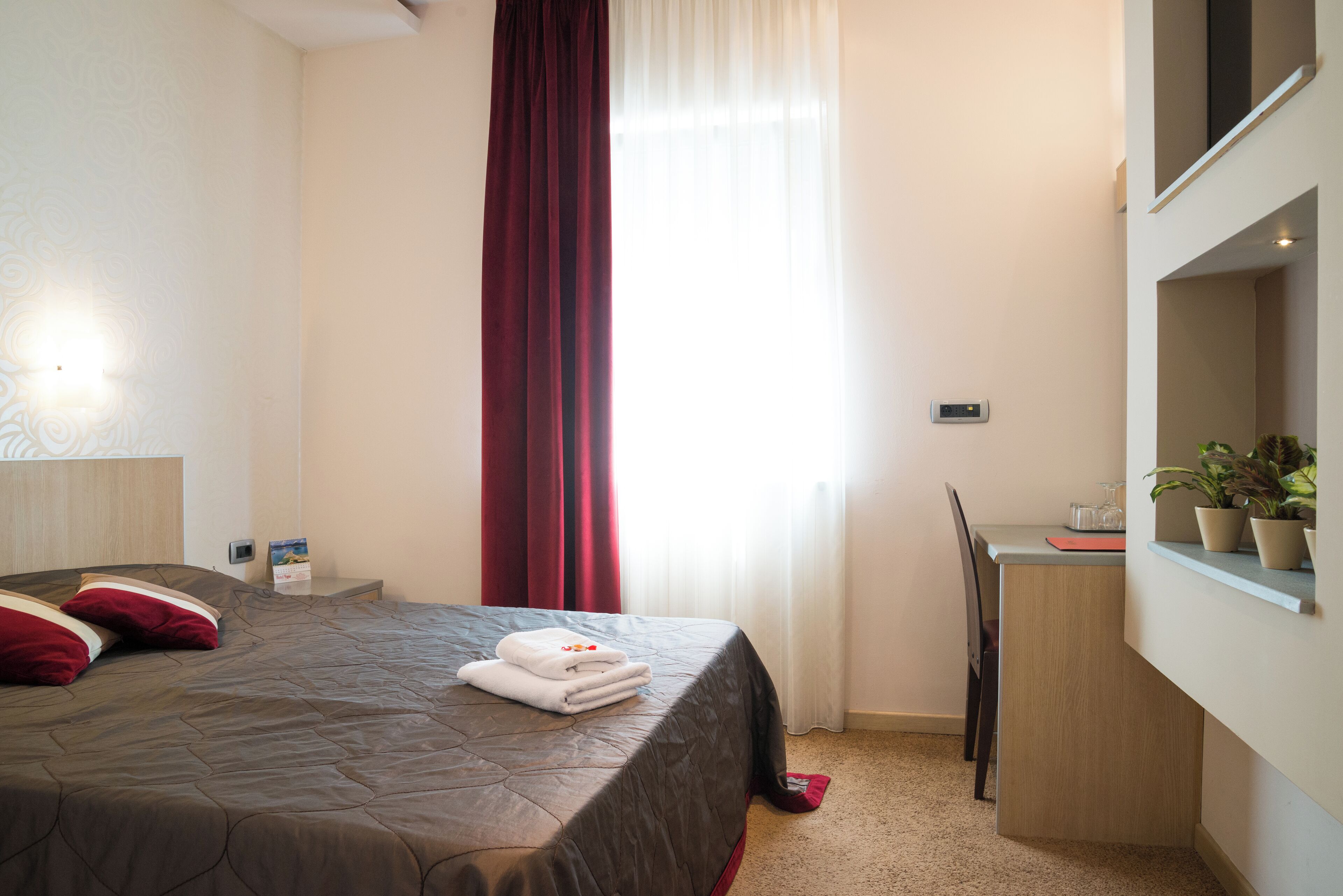 Habitación de hotel con cama, escritorio, silla, planta en maceta y ventana con cortinas.