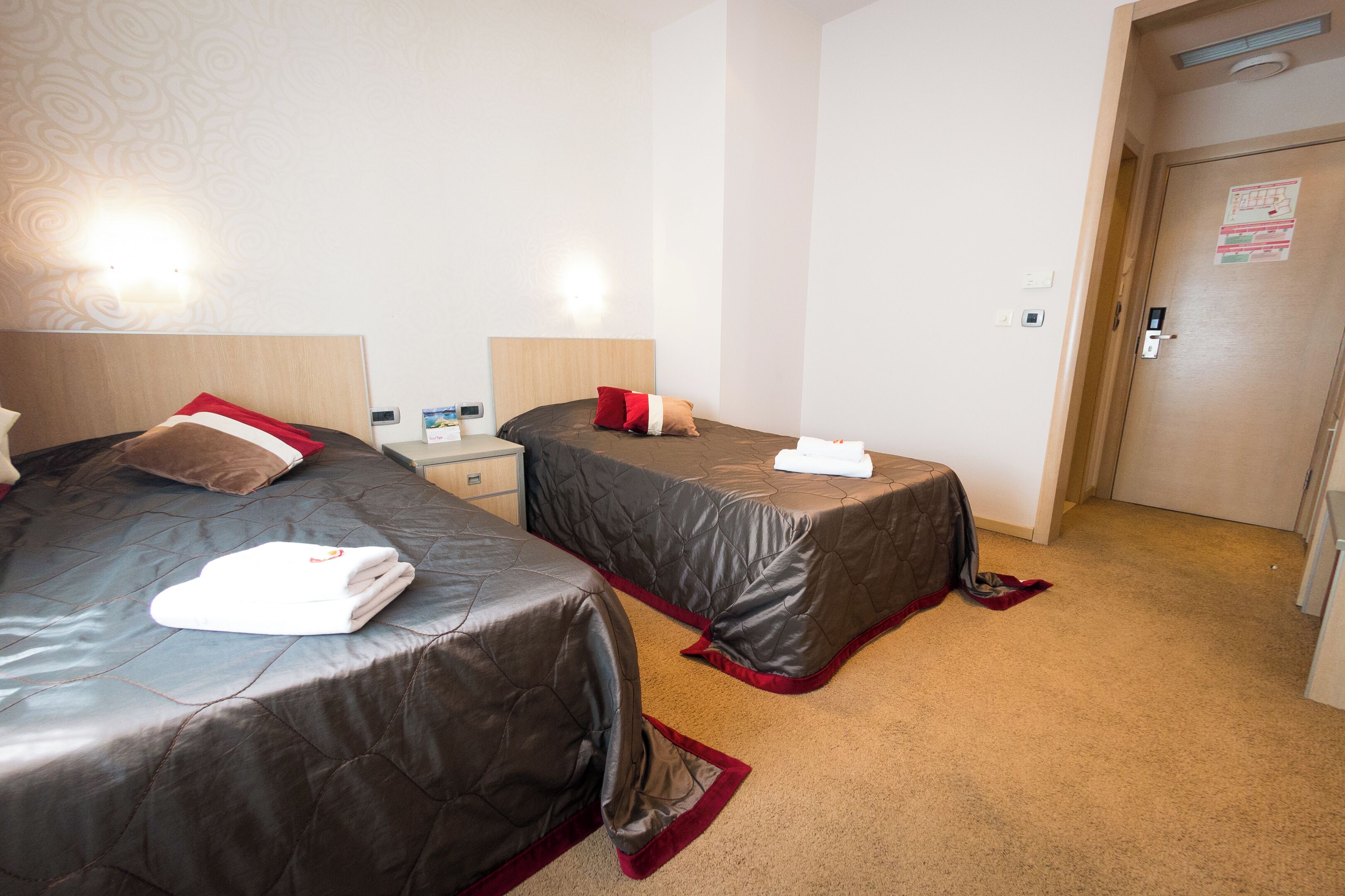 A hotel room with two beds, each with a quilted cover and a red accent pillow. There is a bedside table with a lamp and a small clock. The room has a carpeted floor and a door with a lock and a sign above it.