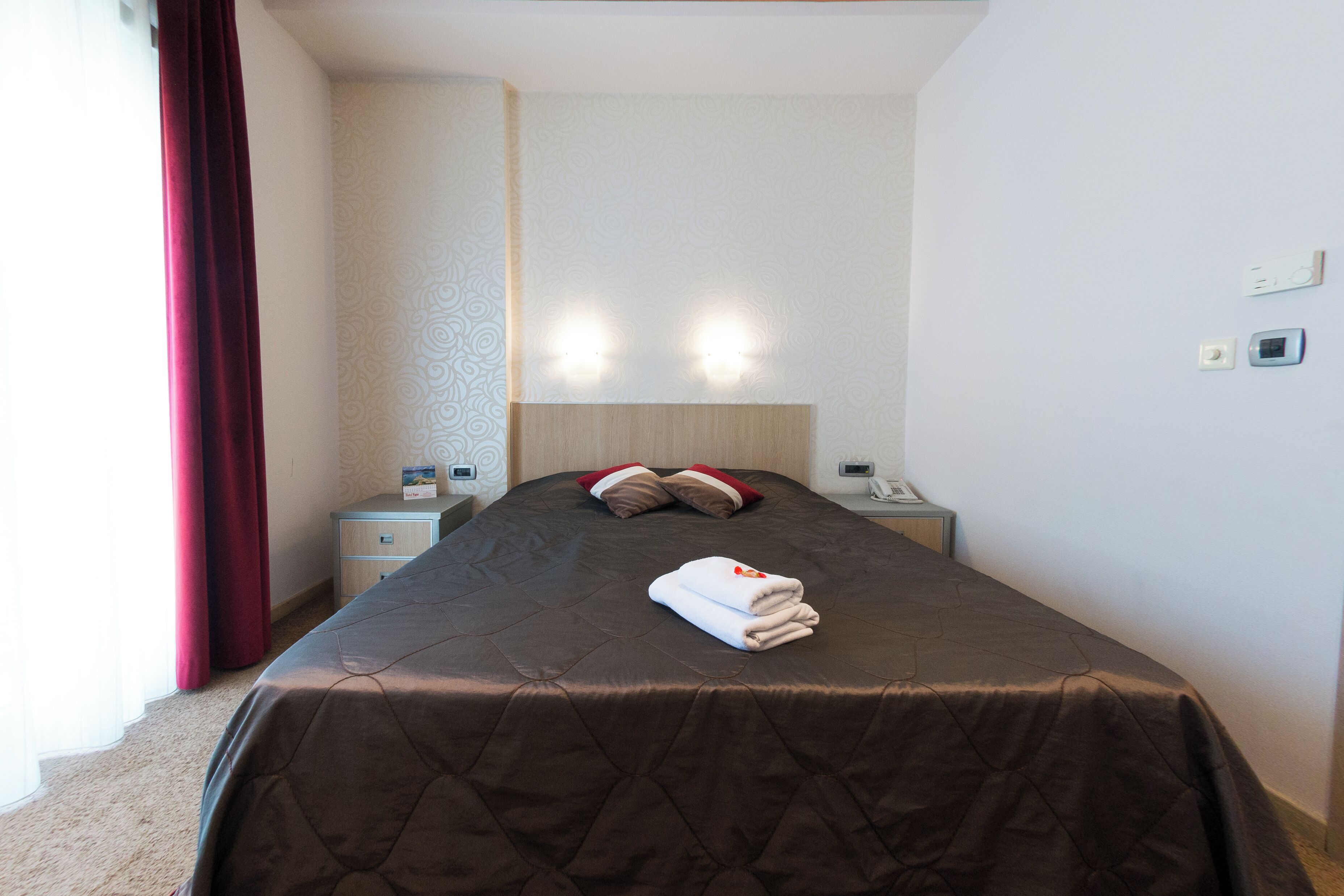 A neatly made bed with a brown quilt, two bedside lamps, a nightstand, and a red curtain.