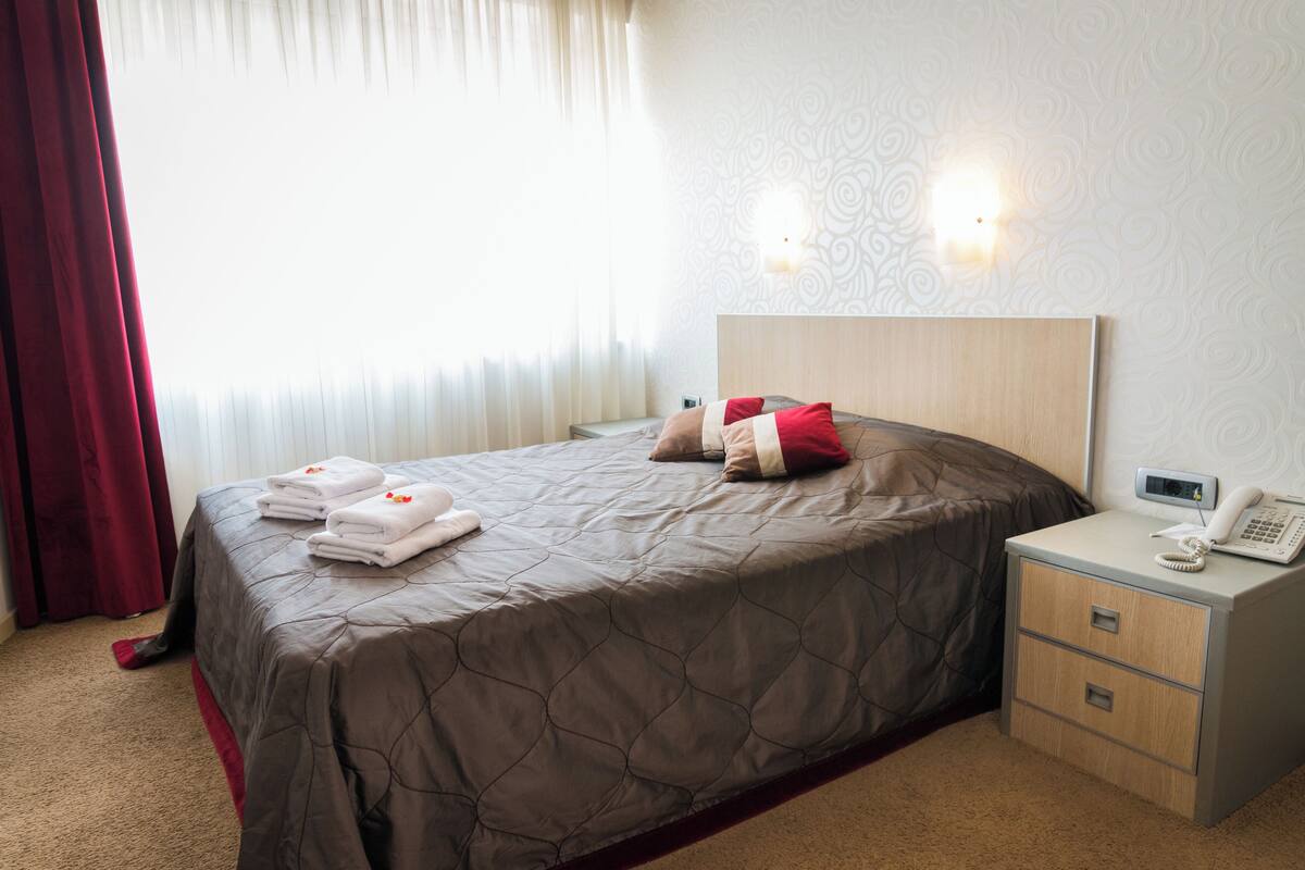 Uma cama bem arrumada com um edredom marrom, uma mesinha de cabeceira com um telefone e um abajur, e uma janela com cortinas finas.