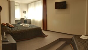 A modern hotel room with a large bed, a desk with a chair, a flat-screen TV mounted on the wall, and a bathroom with a mirror and curtains. - Vigor Hotel (Novi Sad)