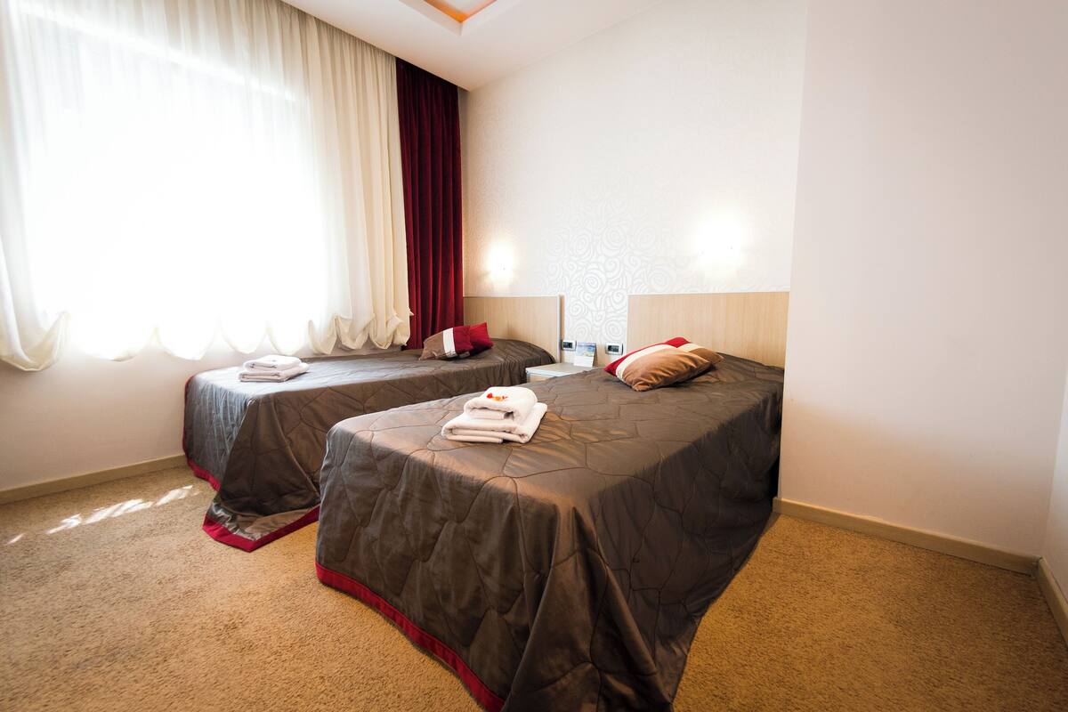 Quarto de hotel com duas camas, cada uma com colcha escura e almofadas vermelhas. Há uma janela com cortinas brancas leves e um tecido vermelho.