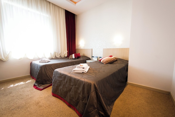 Habitación de hotel con dos camas, cada una con un cubrecolchón oscuro y almohadas rojas. Se ve una ventana con cortinas blancas traslúcidas y un adorno rojo.