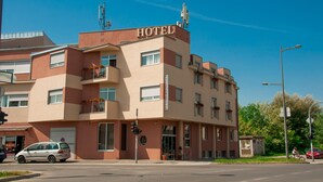 A hotel with a prominent "HOTEL" sign, multiple balconies with flower boxes, and a street view with a parked car and a cyclist. - Vigor Hotel (Novi Sad)