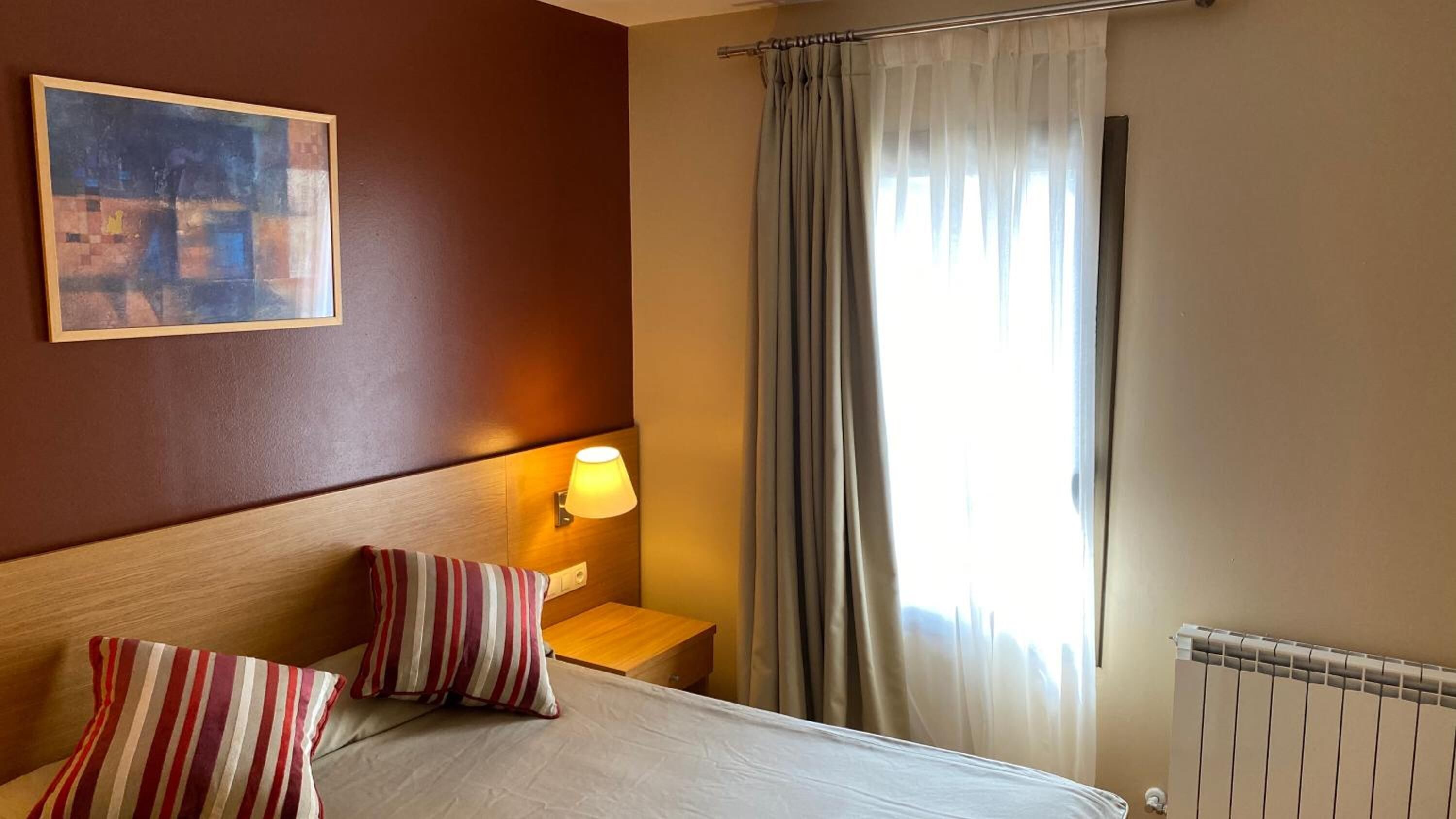 A hotel room with a bed, striped pillows, a nightstand with a lamp, a painting on the wall, and a window with curtains.