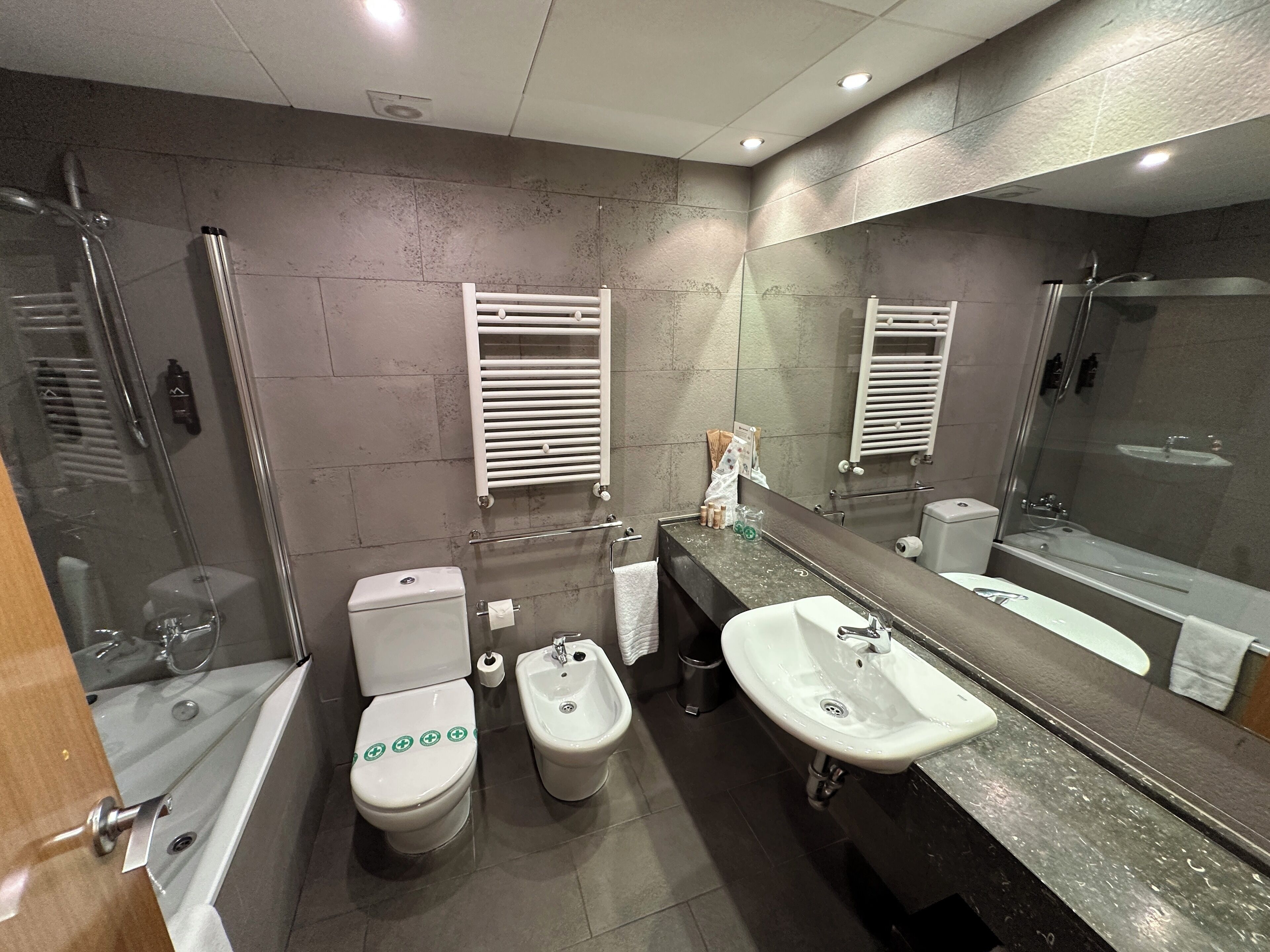 Un baño moderno con ducha de vidrio, dos inodoros, un bidé, un lavamanos y un espejo grande.
