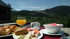 Desayuno con croissants, jamón, queso, sandía y jugo de naranja sobre una mesa con vista panorámica a las montañas.