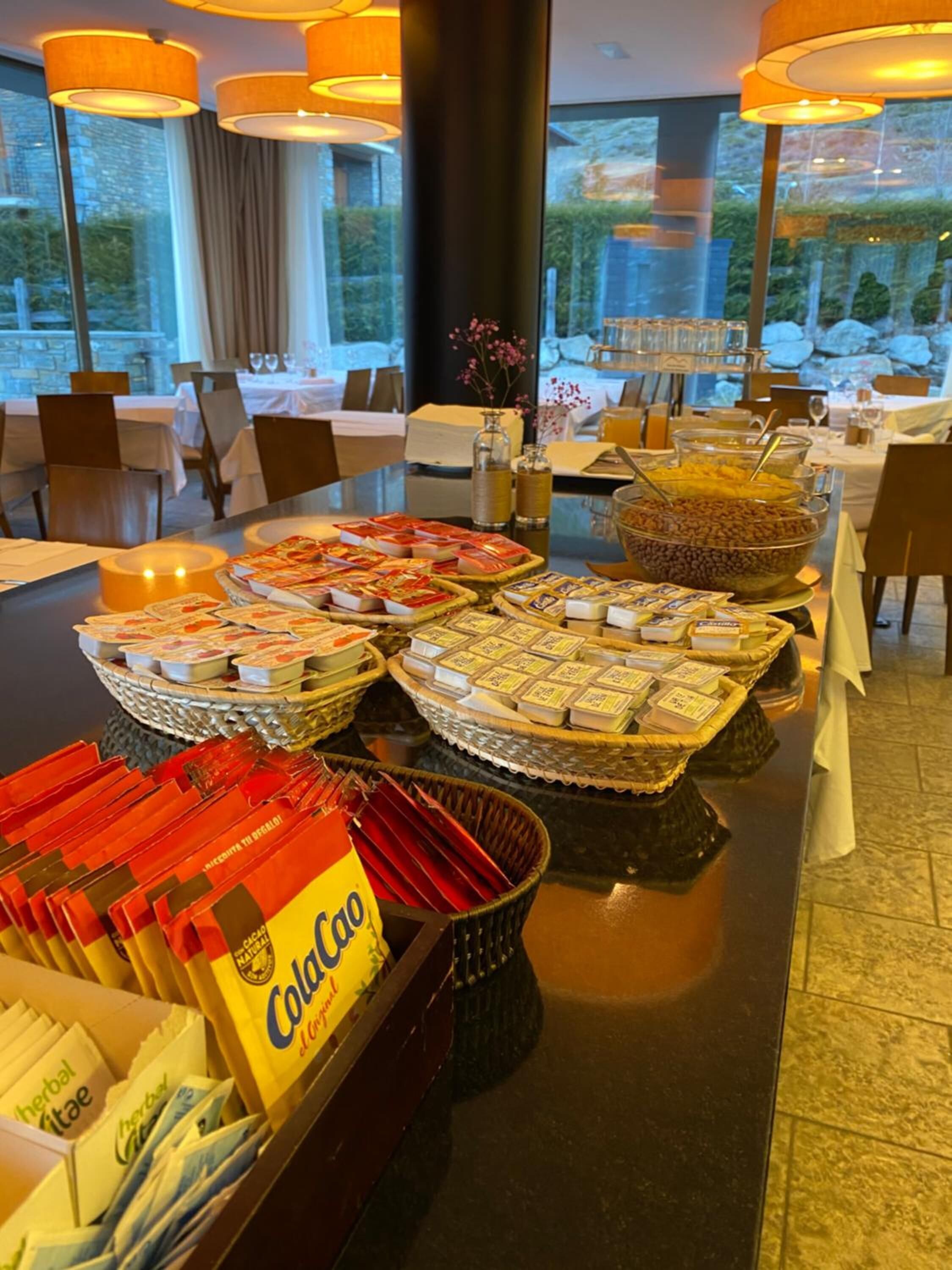 A buffet setup with various food items, including cheese, snacks, and beverages, in a well-lit dining area with large windows.