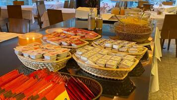 A buffet setup with various food items, including cheese, snacks, and beverages, in a well-lit dining area with large windows.