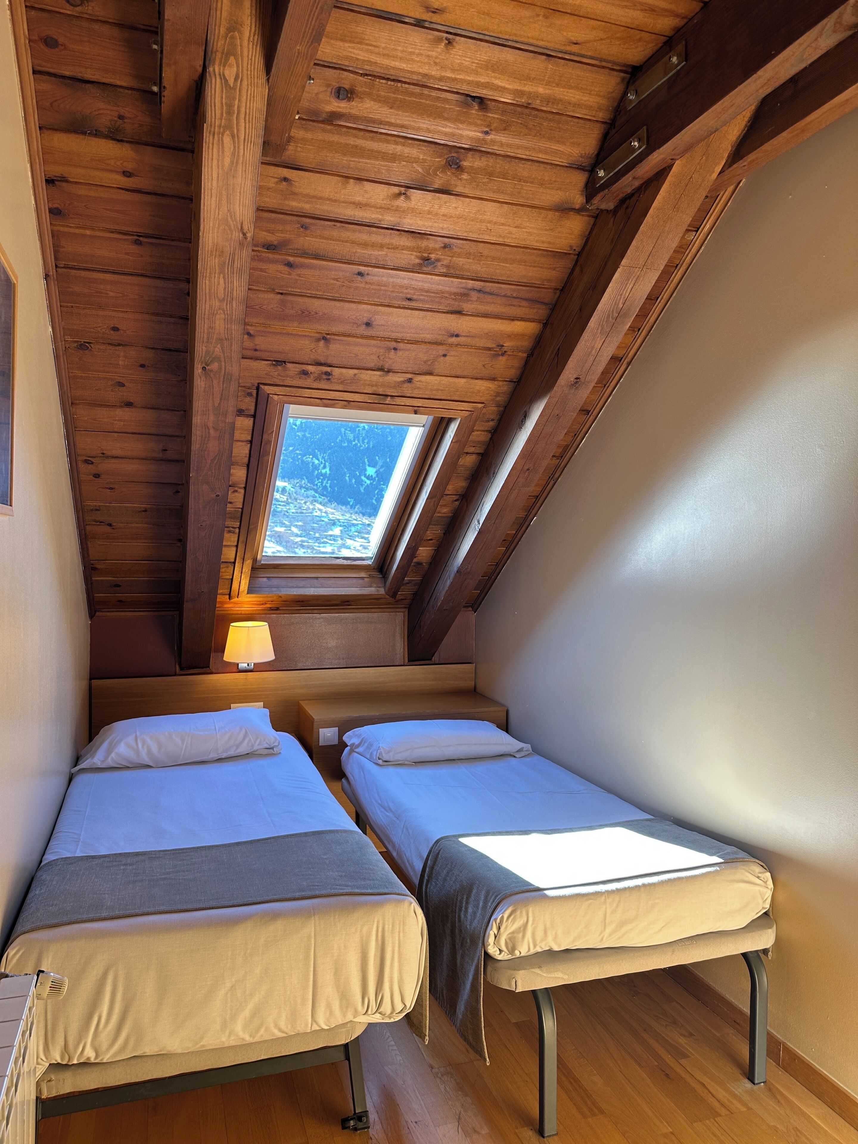 A small, cozy attic bedroom with two single beds, wooden beams, a skylight, and a nightstand with a lamp.