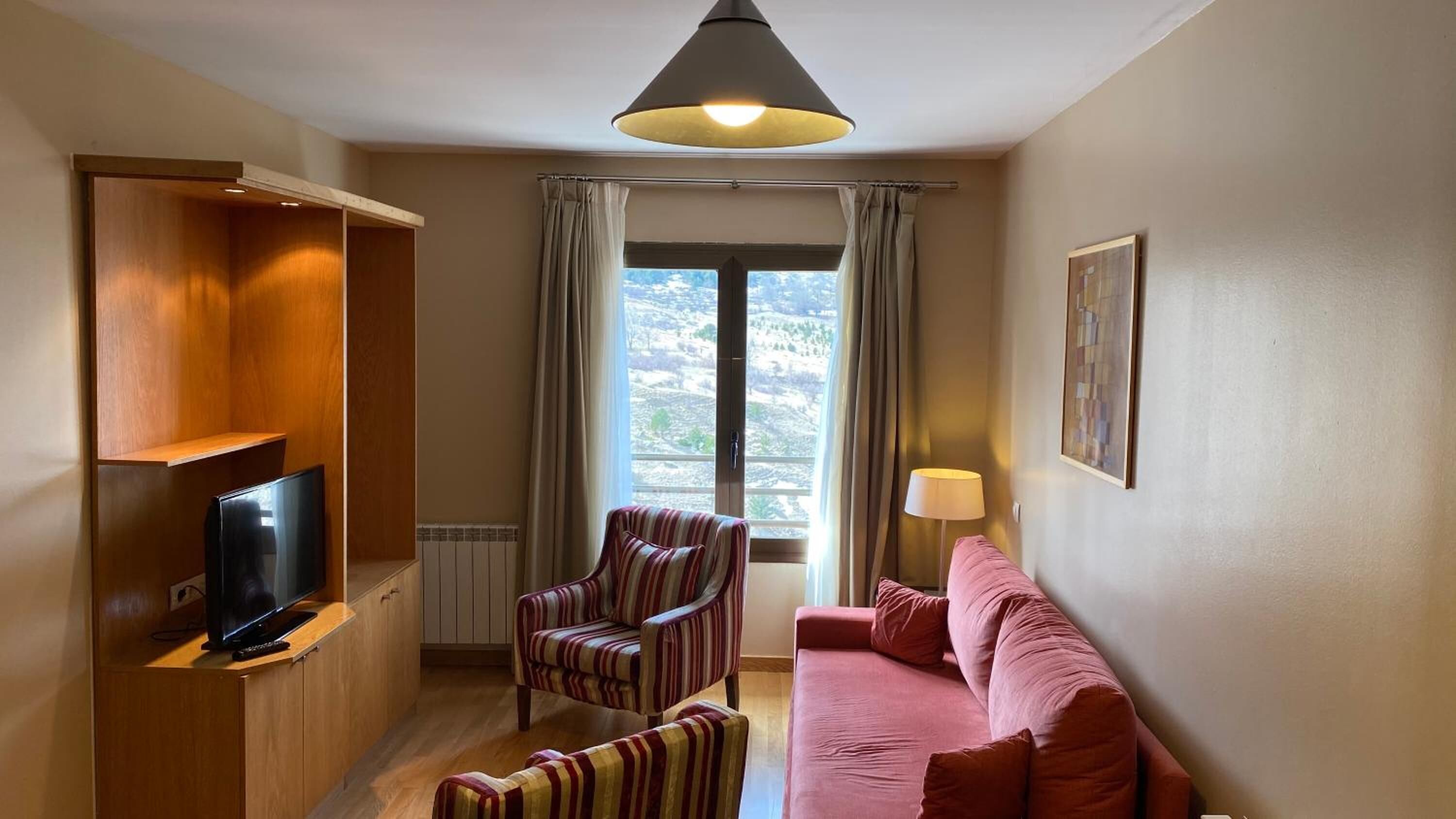 Un espacio habitable pequeño con un televisor en un estante de madera, un sillón a rayas, un sofá rosa y un ventanal con cortinas.