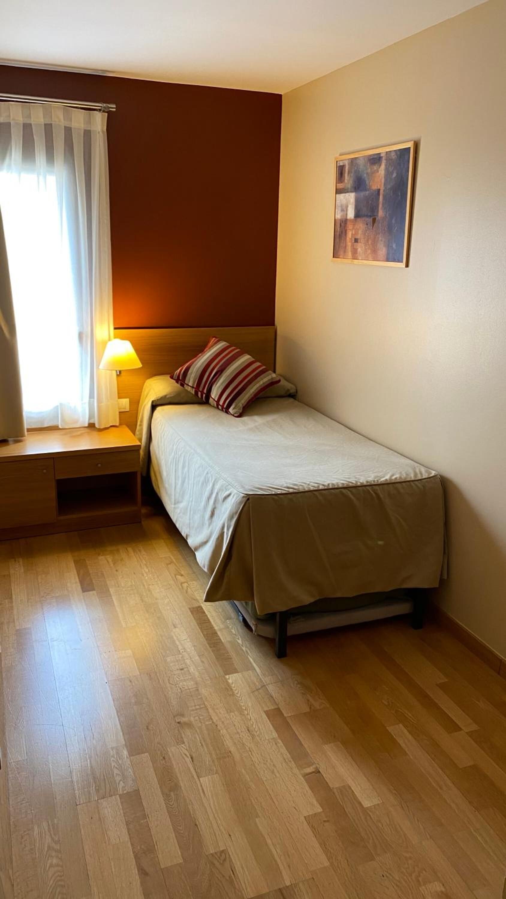 Una cama individual con cabecera, una mesita de noche con lámpara, una ventana con cortinas y un cuadro en la pared.