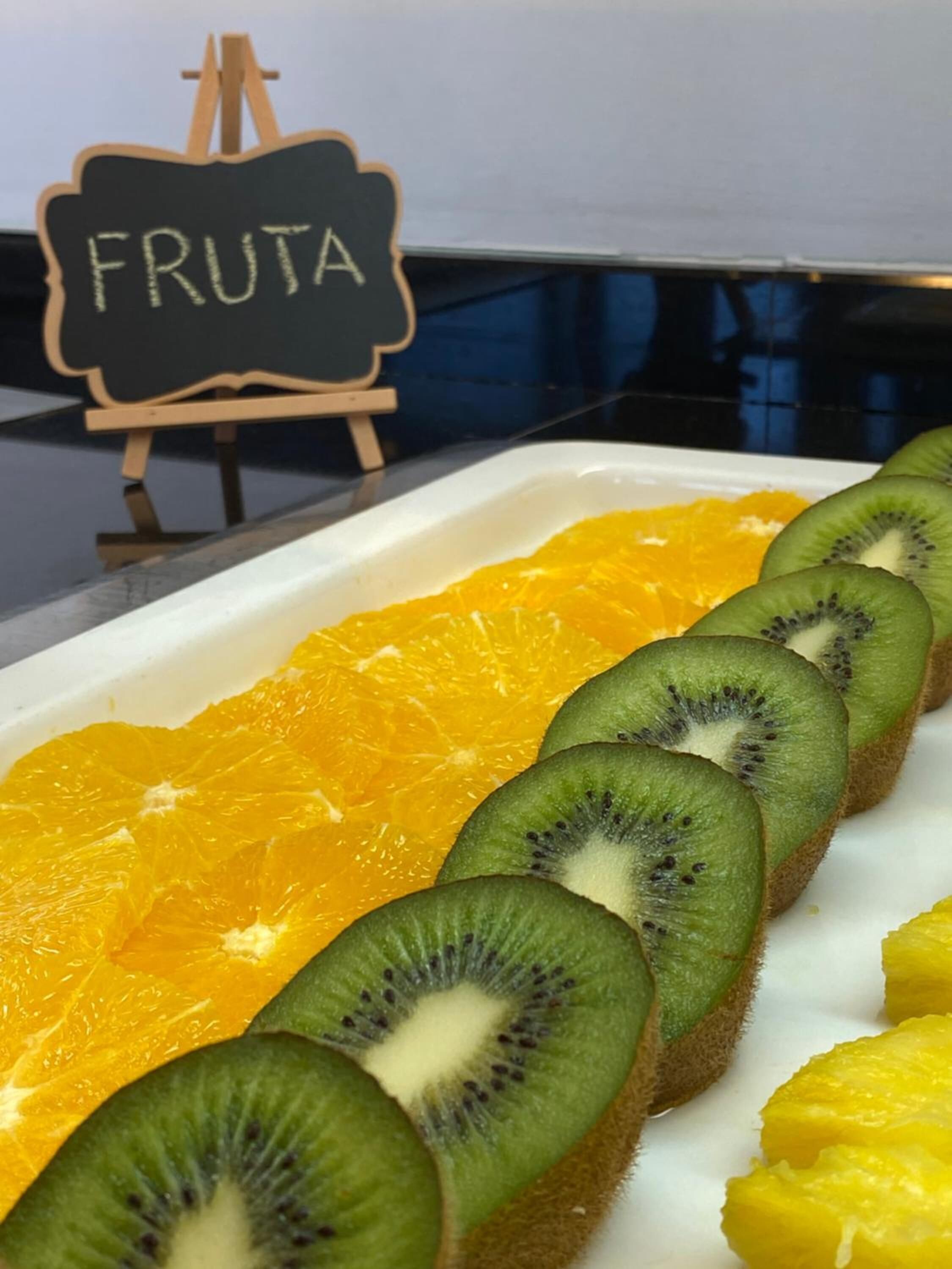 A tray of sliced fruits including oranges and kiwis, with a sign reading "FRUTA" in the background.