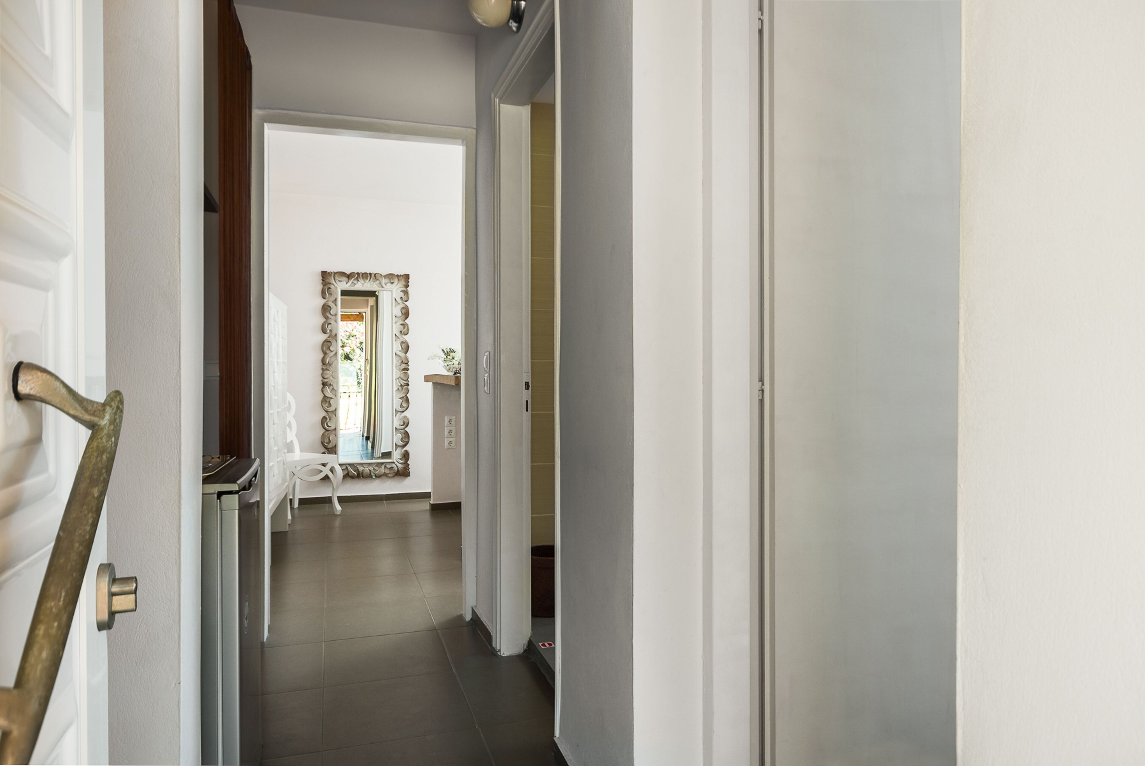 A hallway with a white door on the left, a mirror with a decorative frame, and a white bench in the distance.