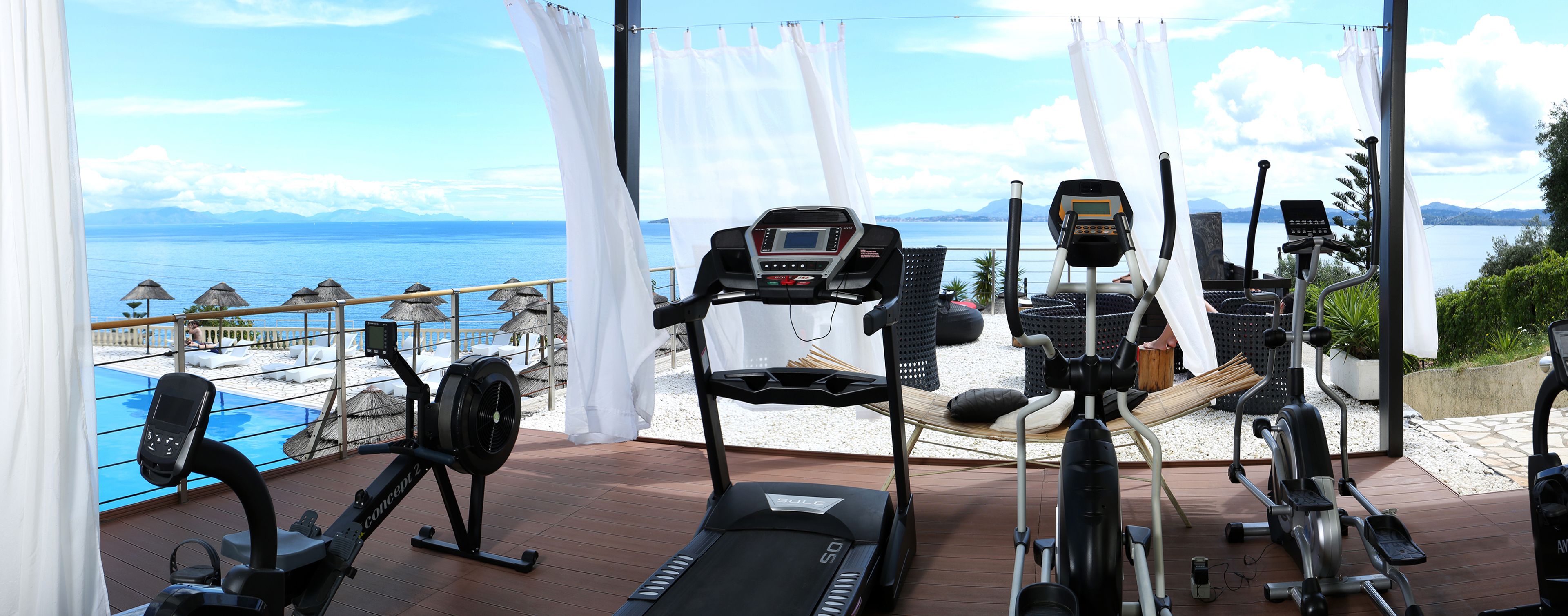 A gym with various exercise equipment, overlooking a beach and the sea.