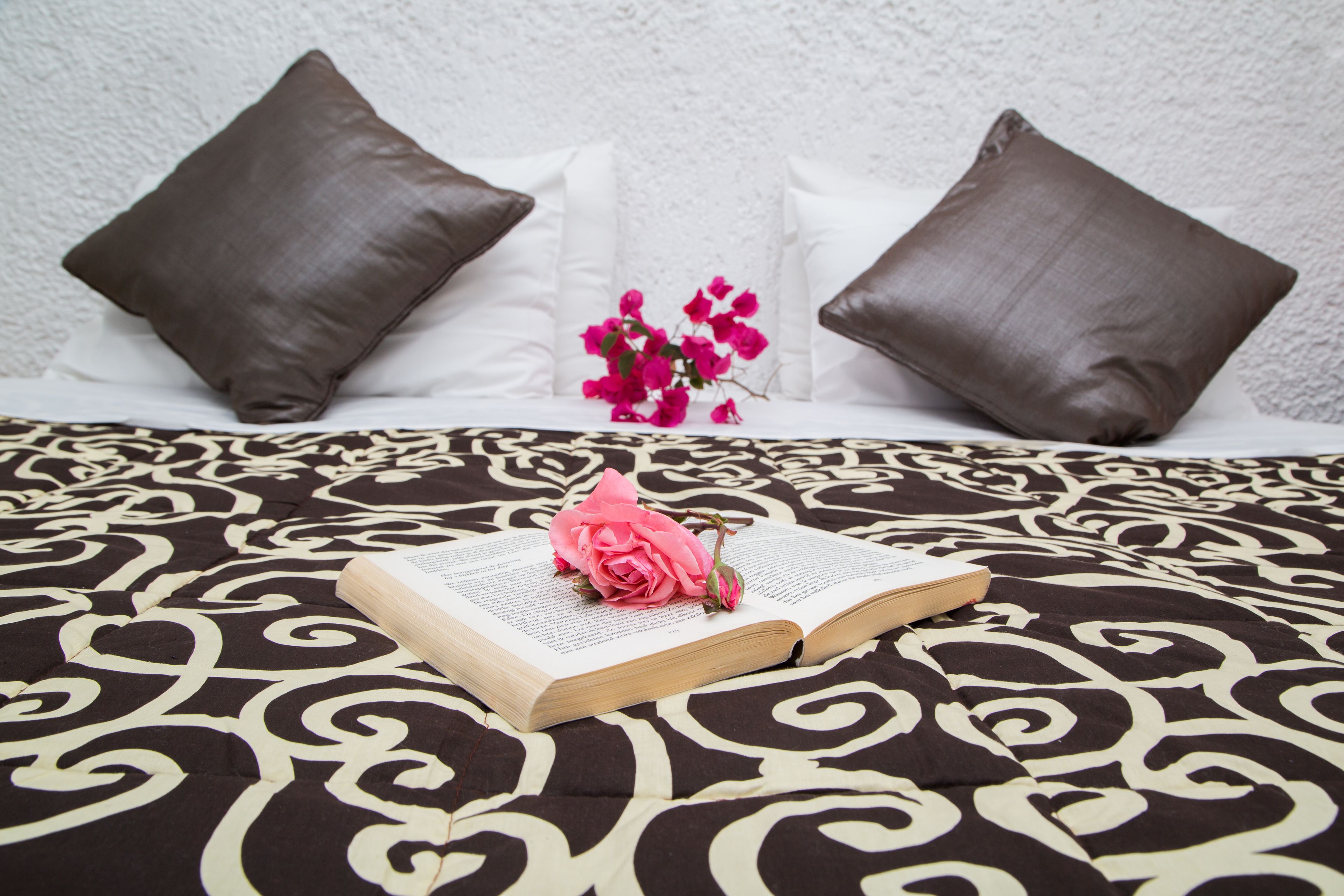 A bed with a patterned bedspread, a book, and a pink flower.