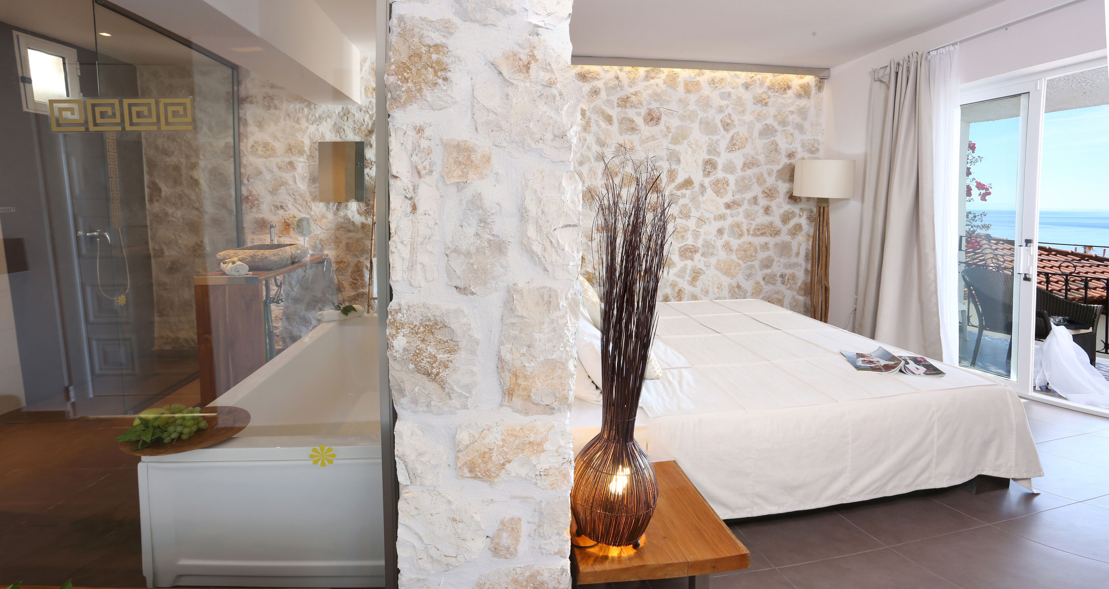 A bedroom with a stone wall, a bed, a bedside table with a vase, and a view of the sea.