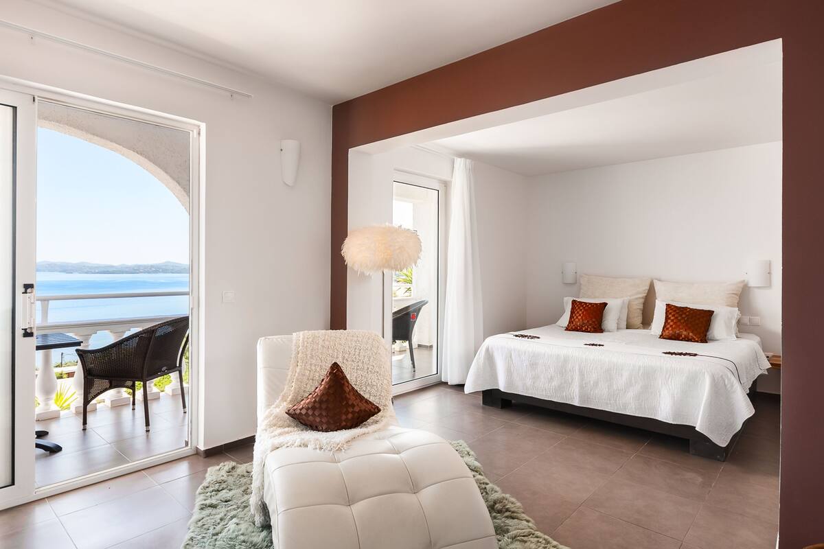 A bedroom with a bed, a chair, a view of the sea, and a balcony.