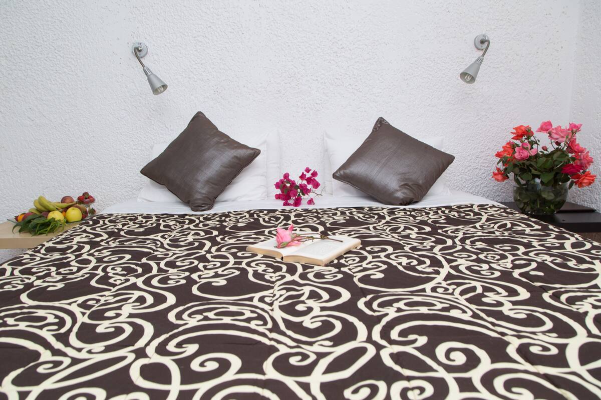 A neatly made bed with a patterned bedspread, two pillows, a bedside table with a vase of flowers, and a fruit basket on the floor.