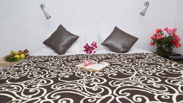 Una cama bien hecha con una colcha estampada, dos almohadas, una mesita de noche con un florero y una cesta de frutas en el suelo.