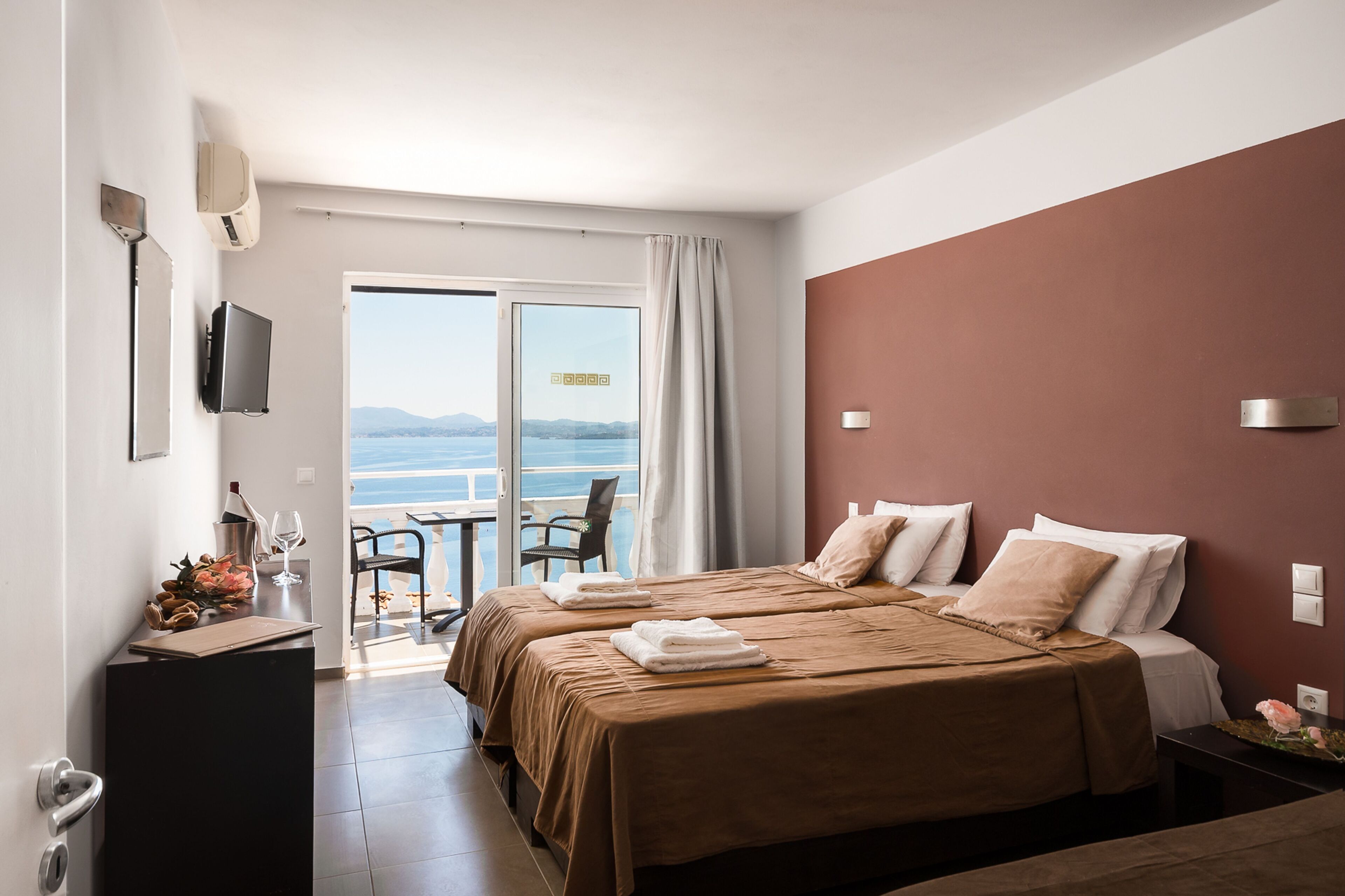 A hotel room with two beds, a TV, a desk with a laptop, and a view of the sea.