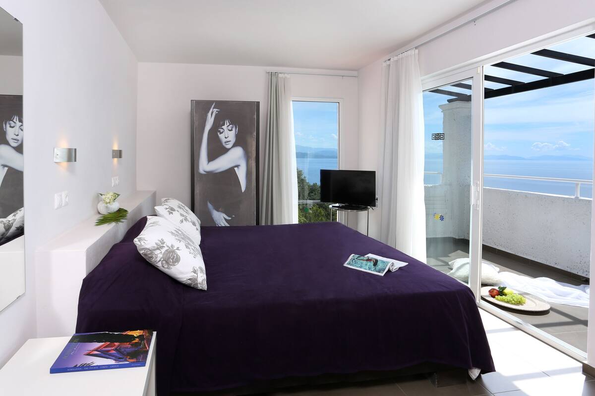 A bedroom with a large bed, a TV, and a balcony with a view.