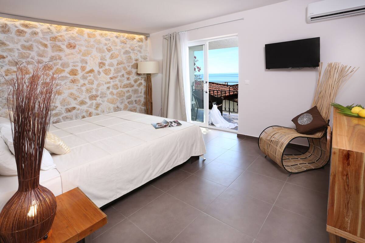 A bedroom with a stone wall, a bed, a TV, a wicker chair, and a view of the sea.
