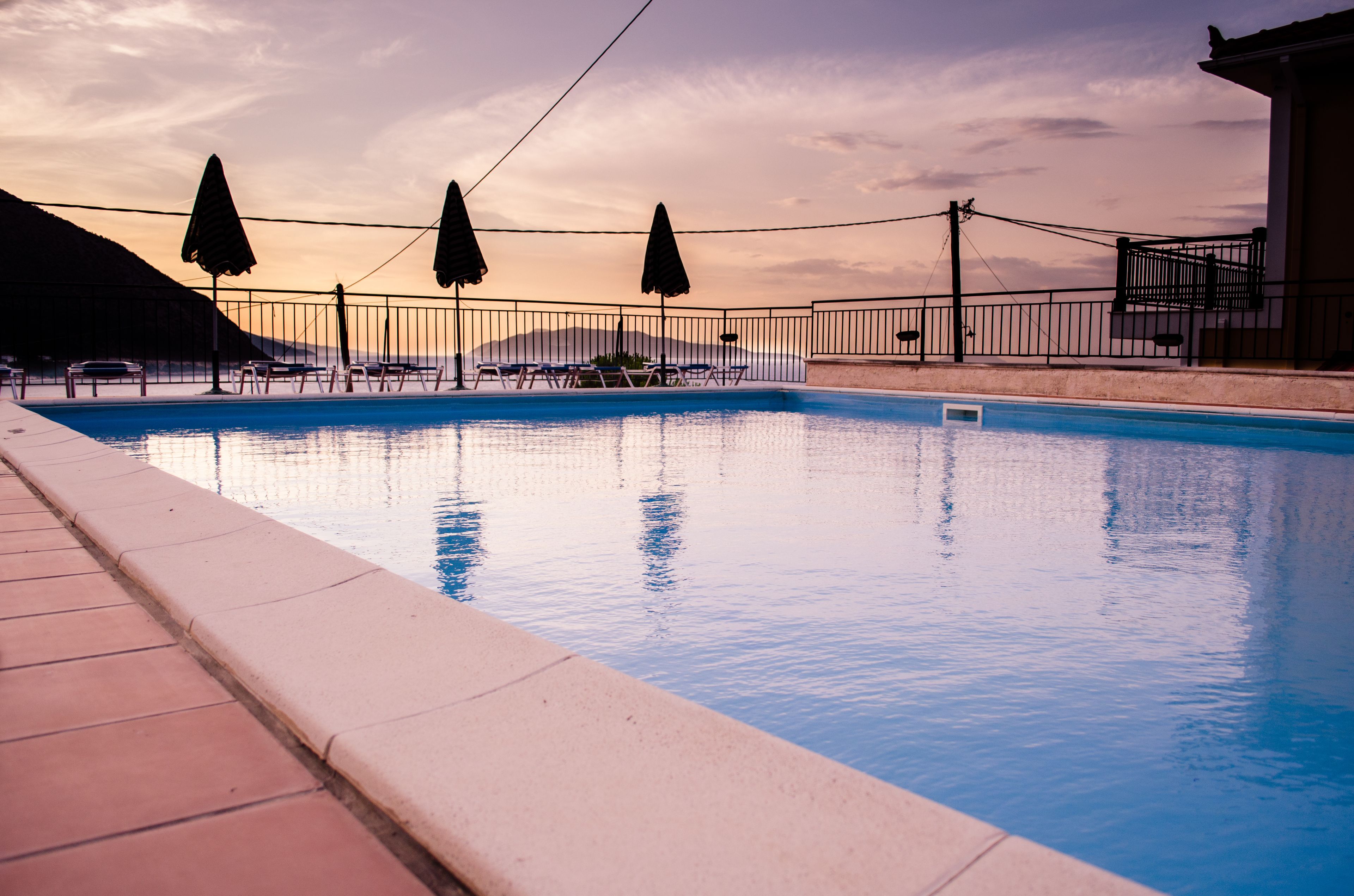 Una piscina con sillas de descanso y sombrillas, rodeada por una valla y un edificio.