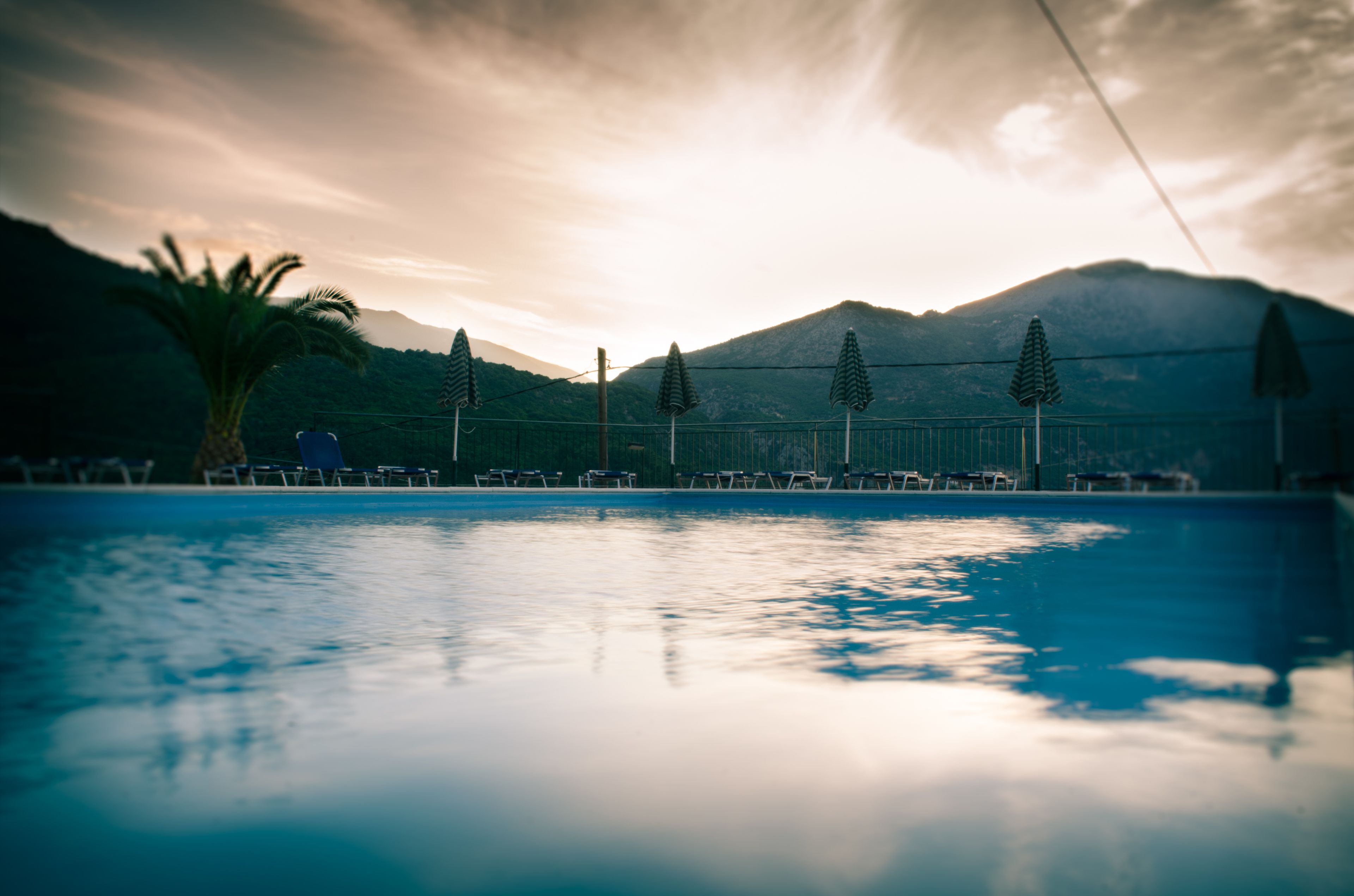 Una piscina con lettini e ombrelloni, circondata da montagne e una palma.