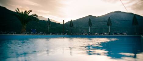 Una piscina con lettini e ombrelloni, circondata da montagne e una palma.