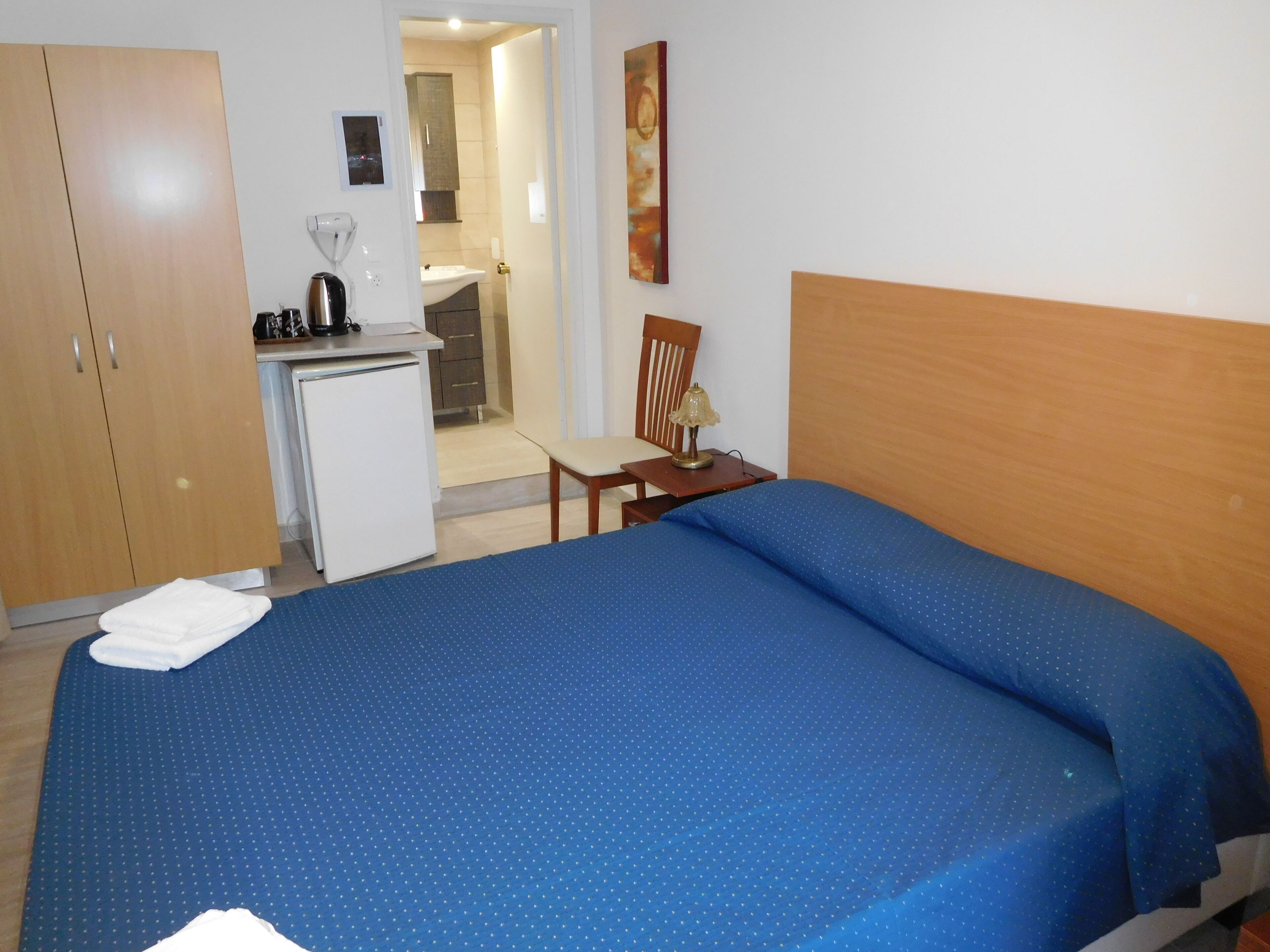A hotel room with a bed, a desk, a chair, a small kitchenette, and a bathroom visible through an open door.