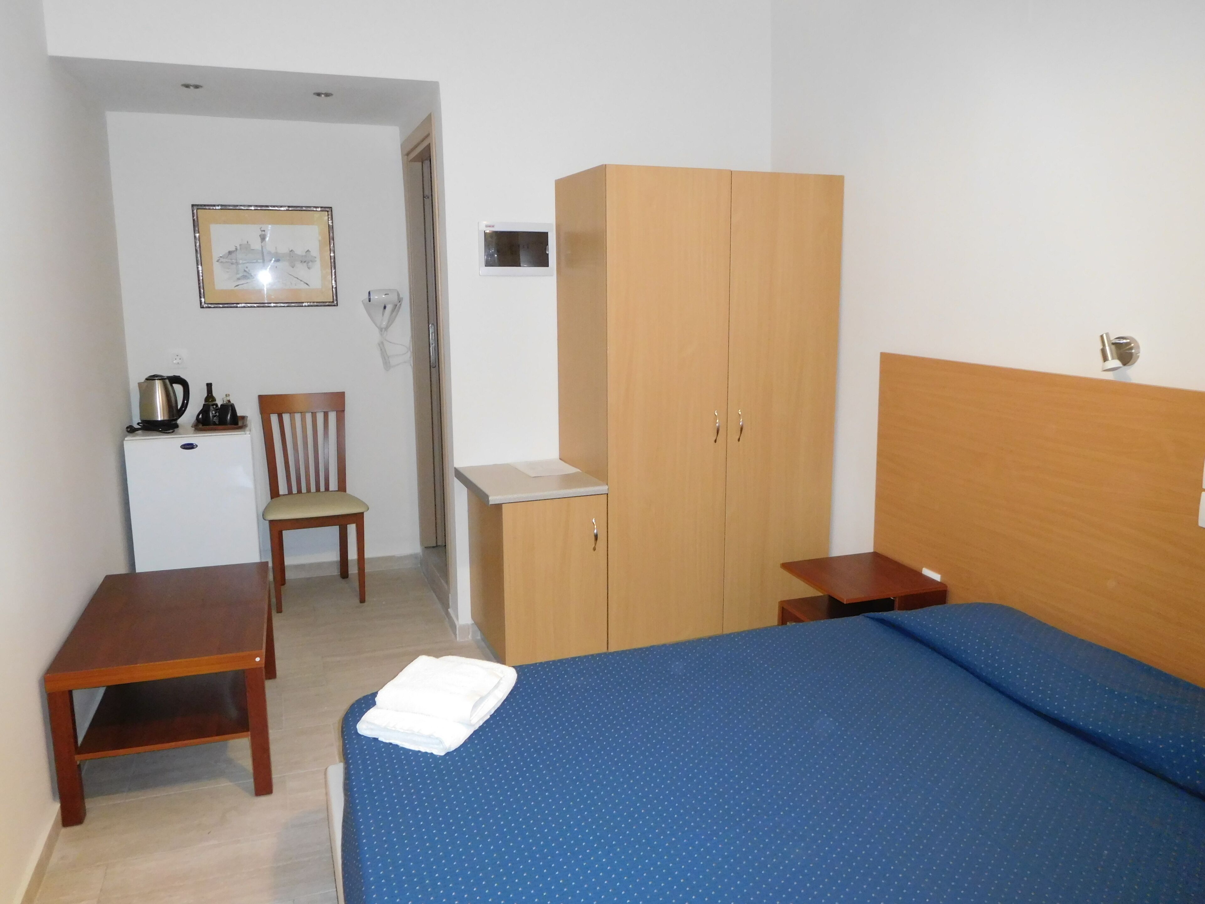 A bedroom with a bed, a wooden wardrobe, a small desk with a chair, and a kitchenette area.