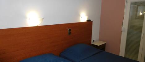 A hotel room with a wooden headboard, two beds with blue bedding, an air conditioning unit, a bedside table with a lamp, and a door with a window.