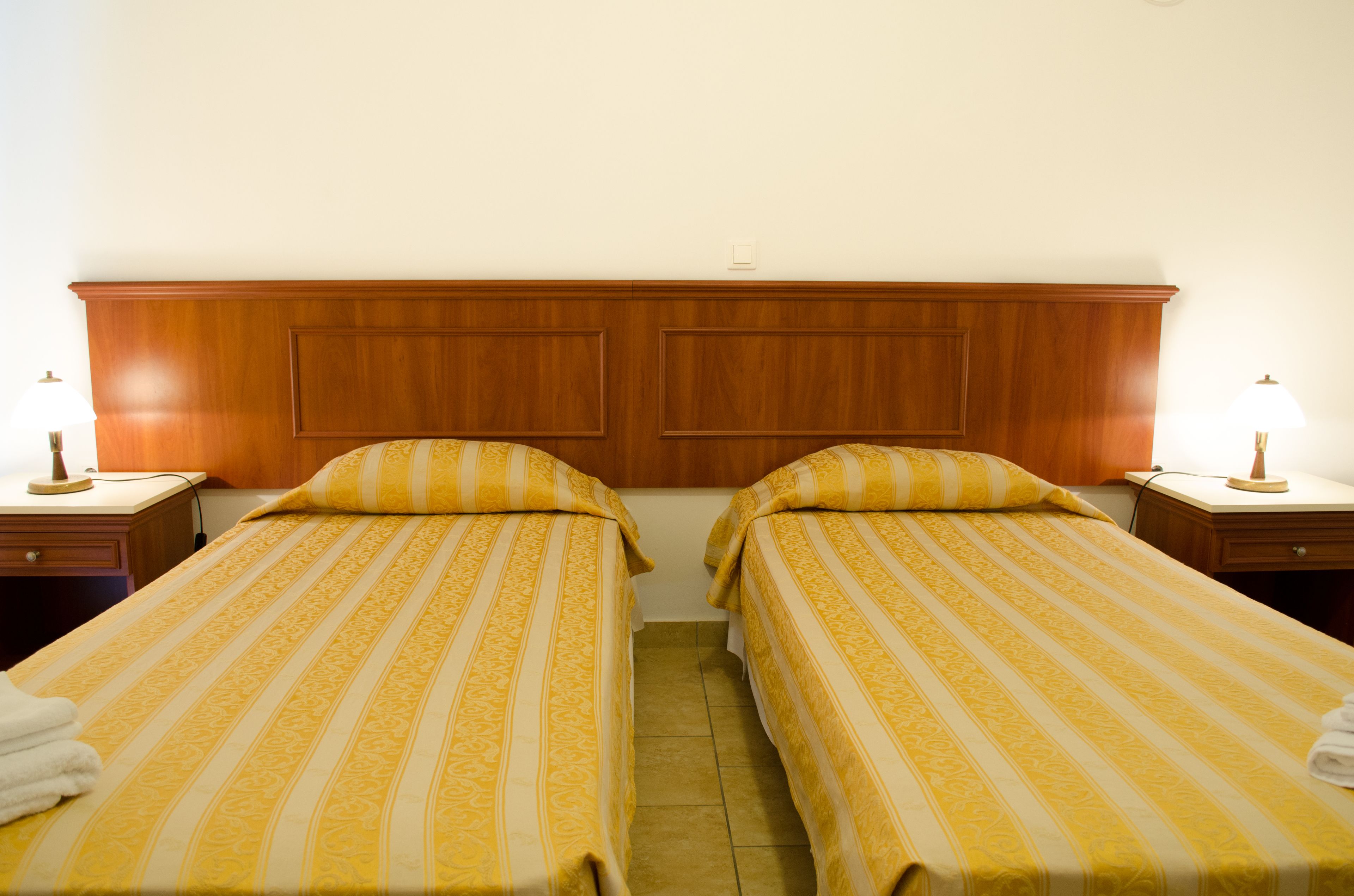 Une chambre d’hôtel avec deux lits simples, une tête de lit en bois, des lampes de chevet et un sol carrelé.