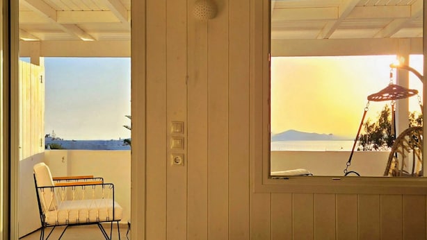 A bedroom with a bed, a chair, and a view of the sea. - Afrodite Boutique Hotel (Paros)