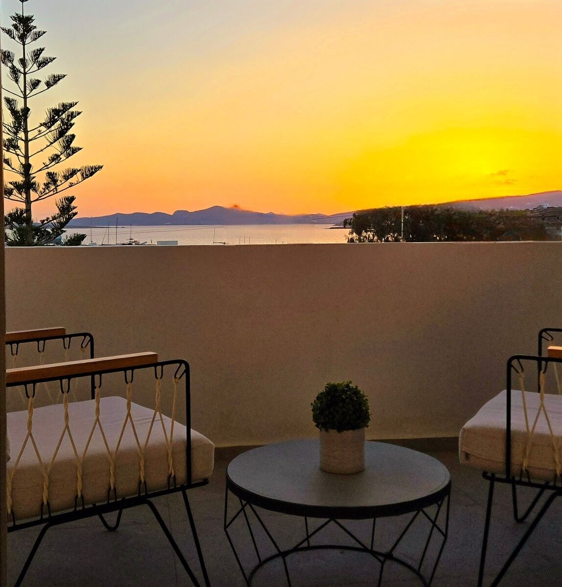 A terrace with two cushioned chairs, a small round table with a potted plant, and a view of a sunset over a body of water with distant mountains.