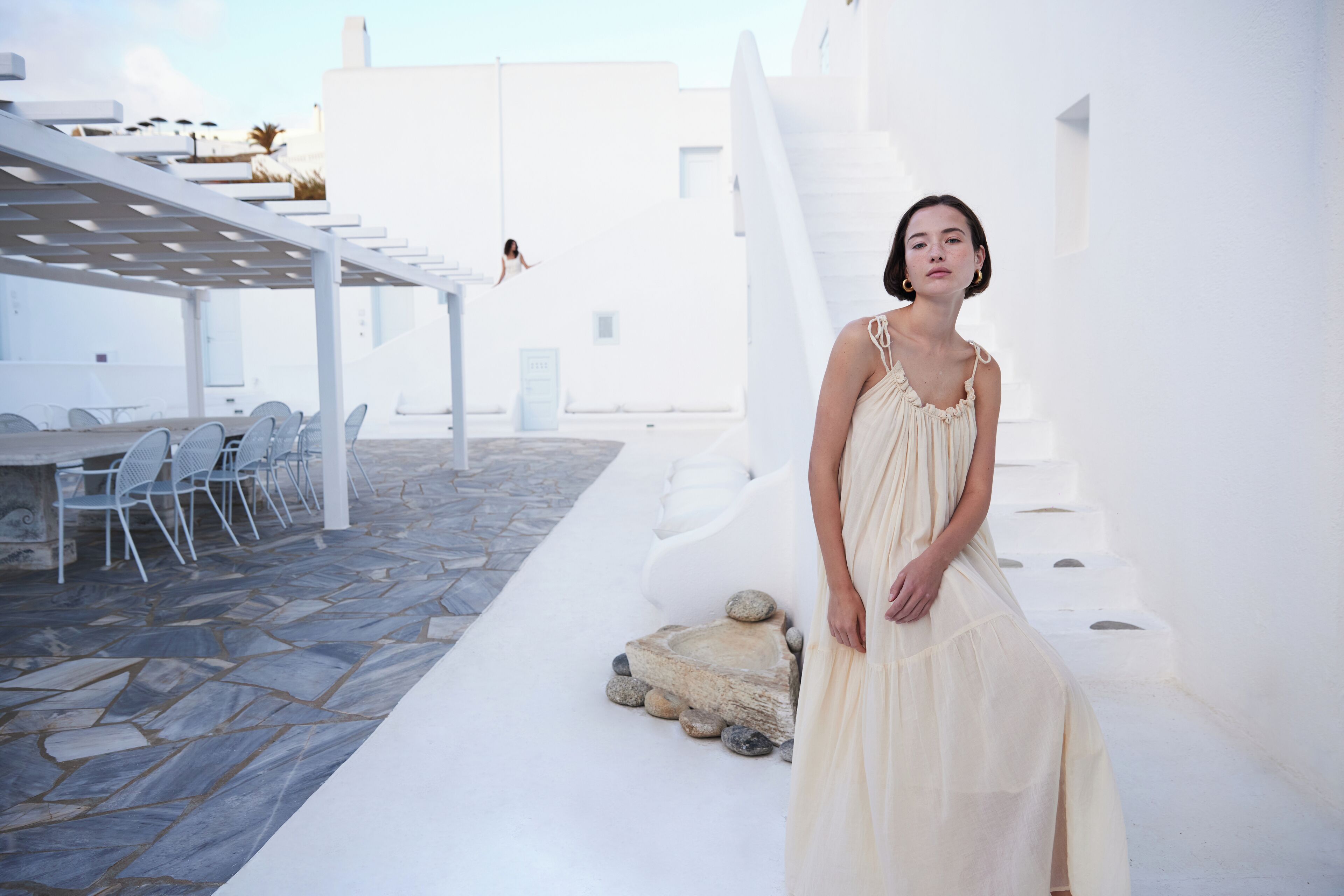 Una mujer con vestido blanco parada en un espacio exterior con paredes blancas, suelo de piedra y una escalera blanca.