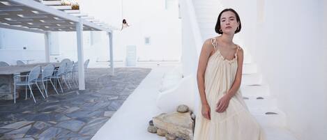 Una mujer con vestido blanco parada en un espacio exterior con paredes blancas, suelo de piedra y una escalera blanca.