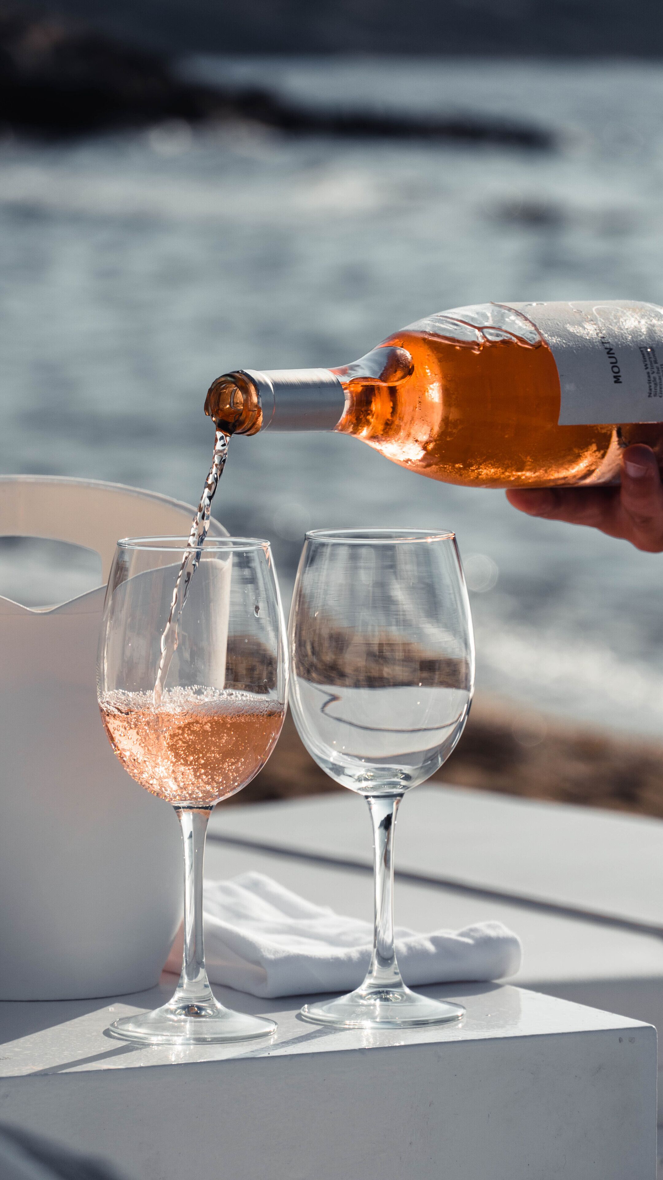 A bottle of rosé wine being poured into two glasses on a table by the sea.