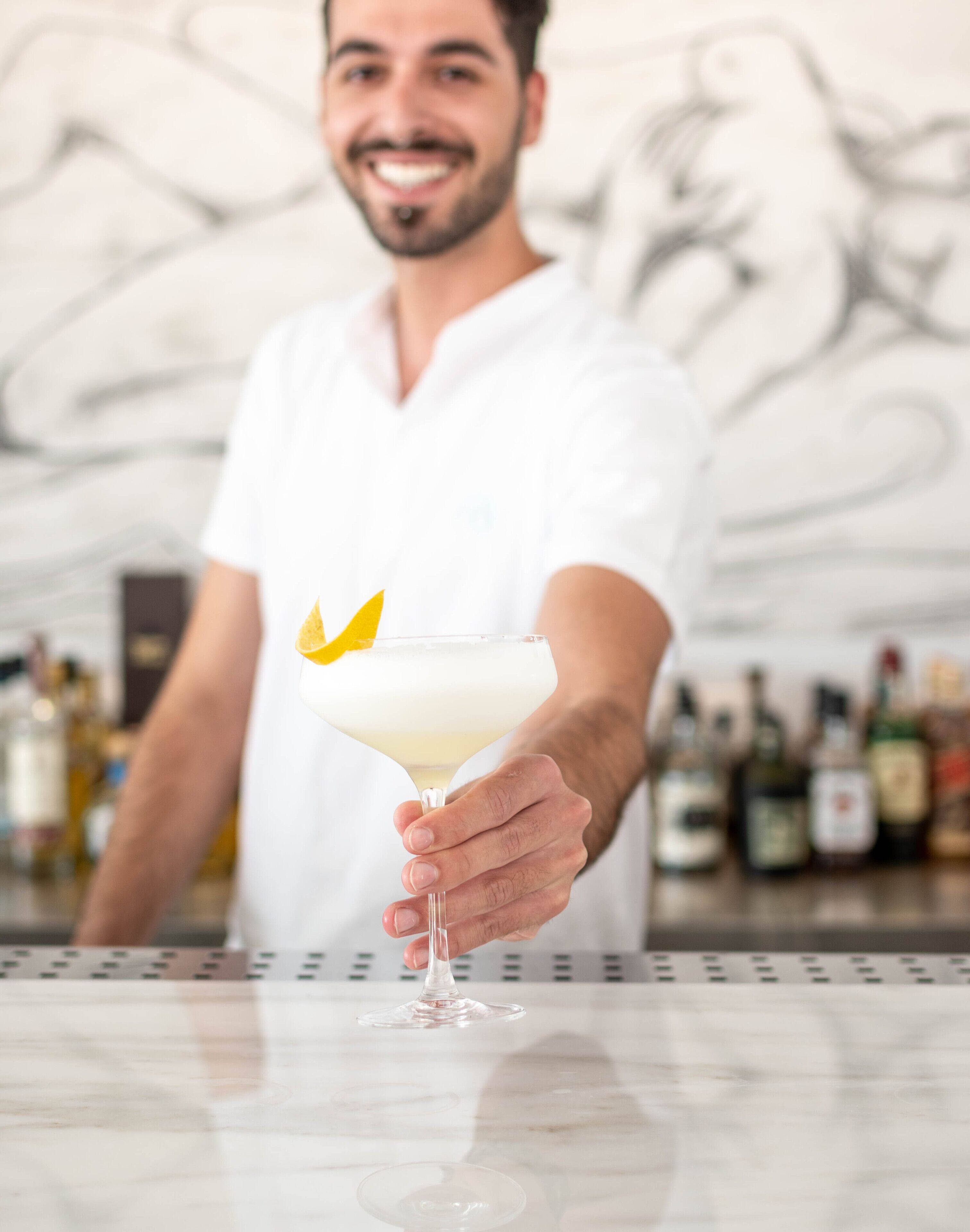 Une personne tenant un verre à martini, décoré d’une feuille jaune, dans un bar où l’on aperçoit des bouteilles en arrière-plan.