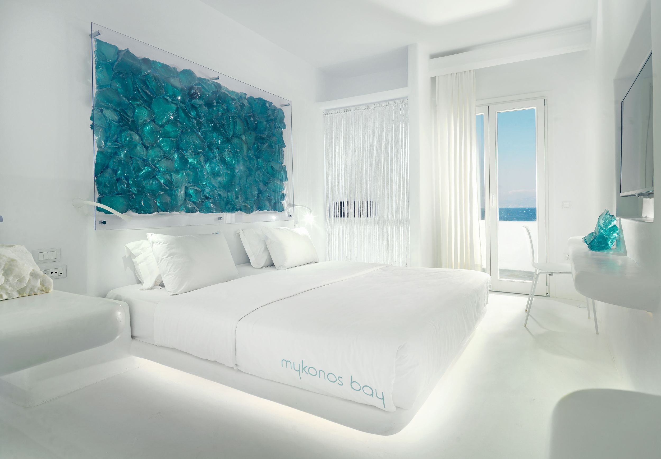 Una habitación de hotel moderna con una cama grande, una obra de arte de cristal sobre la cama, vista al mar por la ventana y una decoración en tonos blancos.