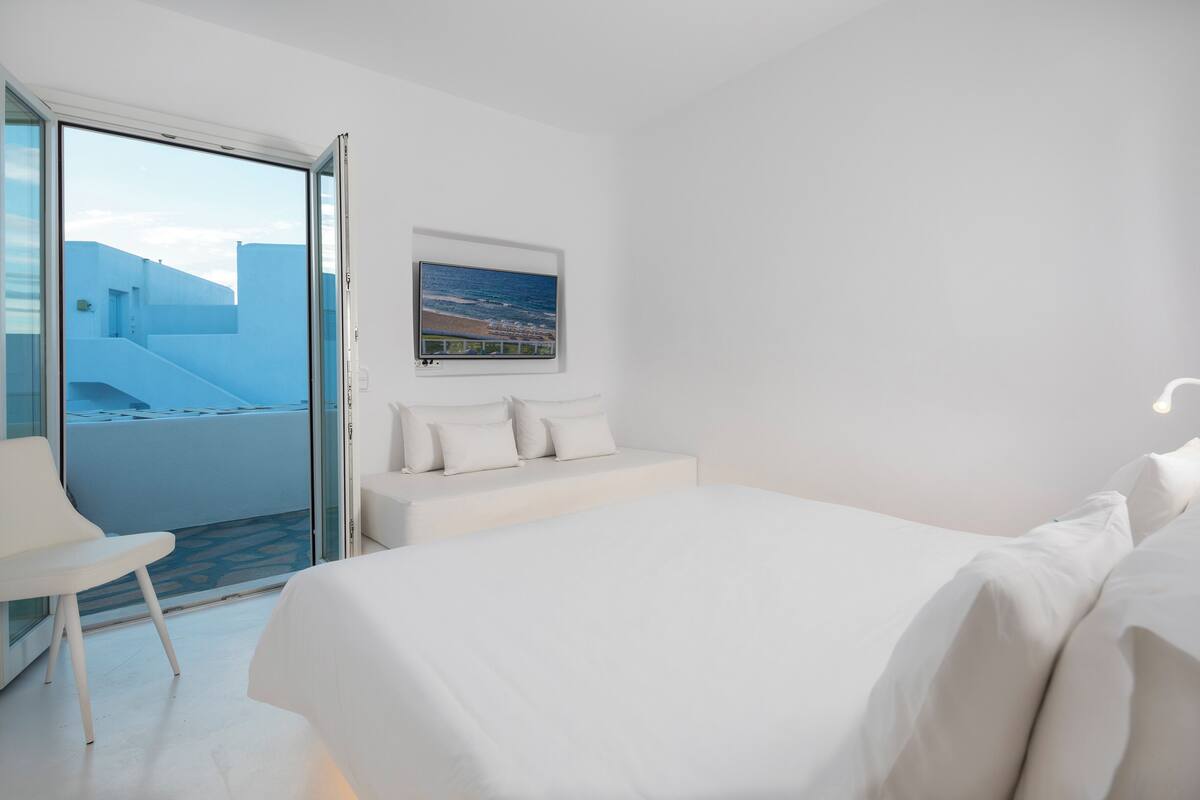 Une chambre d’hôtel moderne avec un grand lit, un canapé, une porte vitrée donnant sur un balcon et un tableau encadré au mur.