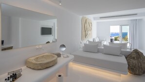 A modern hotel room with a large mirror, a stone sink, a flat-screen TV, a large window with a view of the sea, and a cozy bed with white bedding.