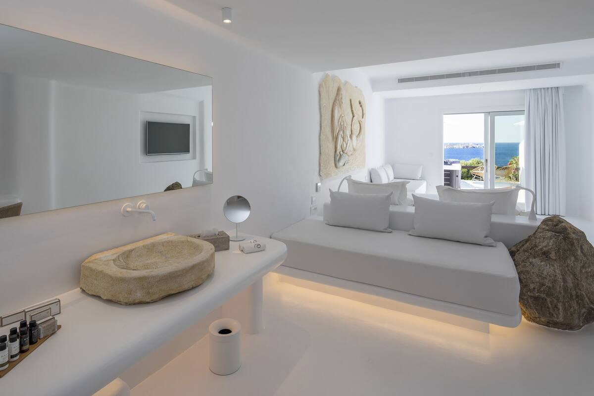 Une chambre d’hôtel moderne dotée d’un grand miroir, d’un lavabo en pierre, d’un téléviseur à écran plat, d’une grande fenêtre donnant sur la mer et d’un lit confortable avec une literie blanche.
