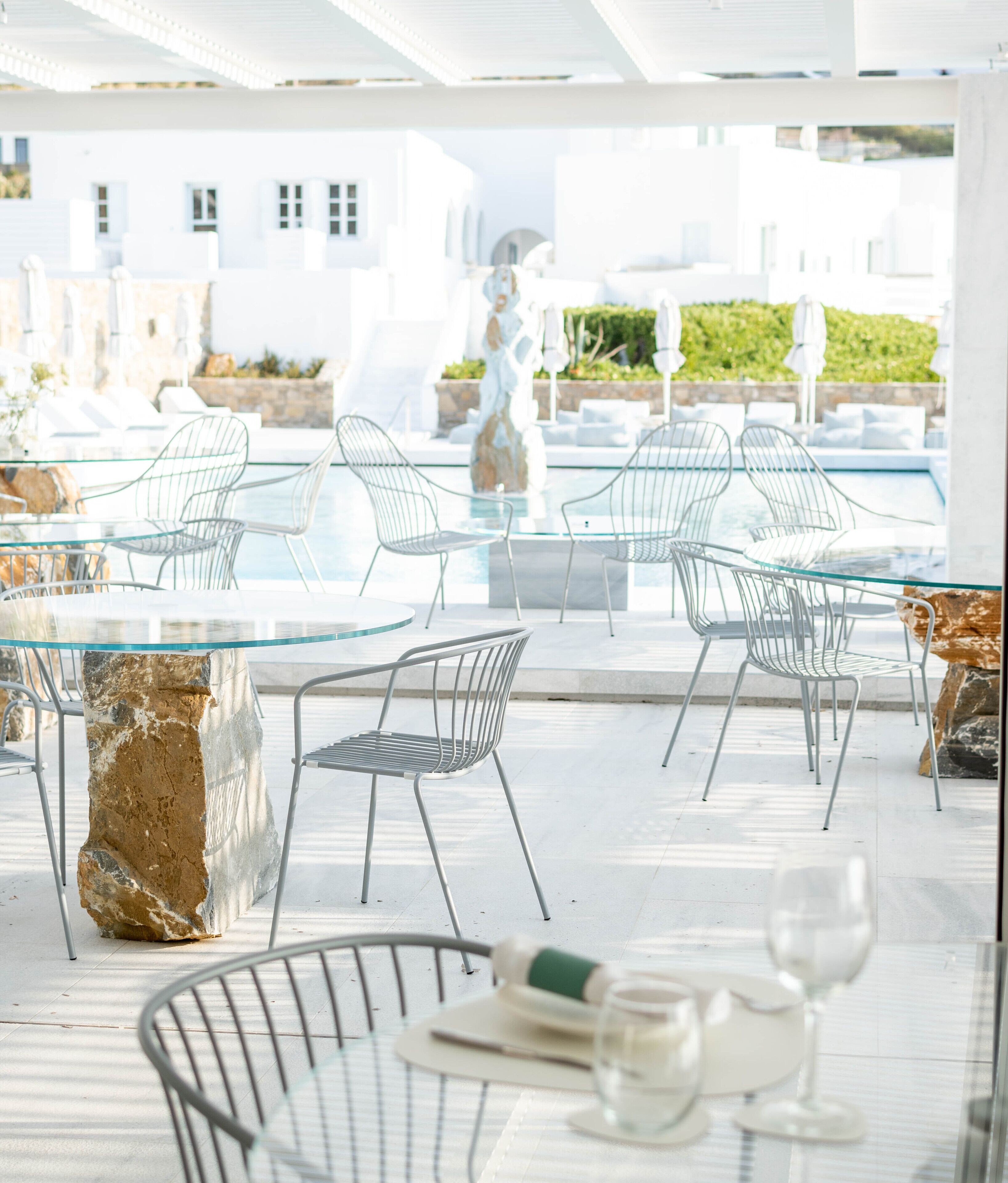 Zona pranzo esterna moderna con tavoli in vetro e sedie in metallo, con vista su una piscina e un edificio bianco.