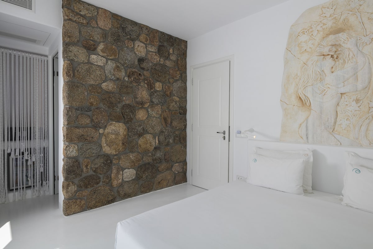Une chambre avec un mur en pierre, un lit recouvert de linge de maison blanc et une grande œuvre d’art en marbre accrochée au mur.