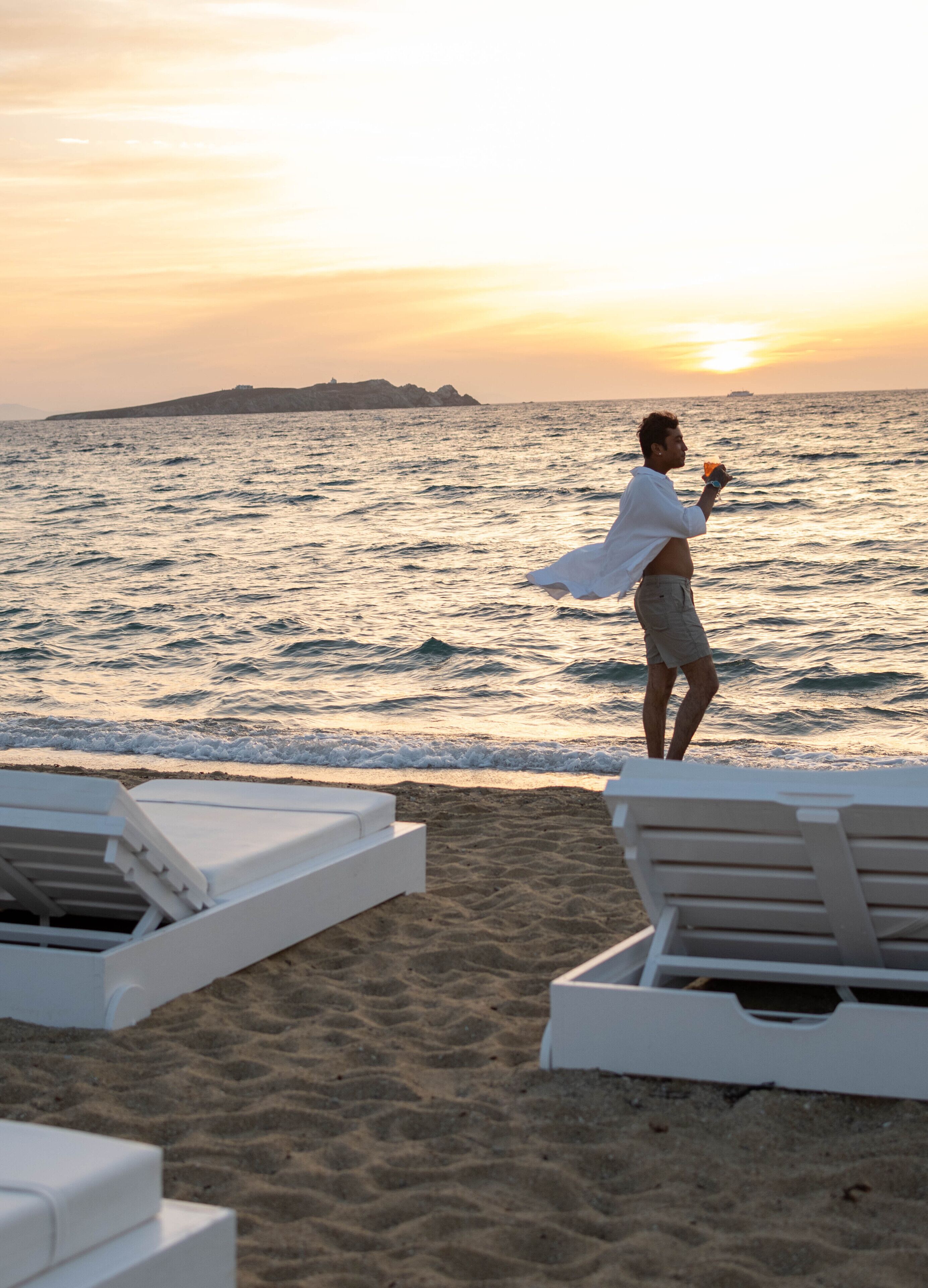 Escena de playa con una persona sosteniendo una bebida, sillas de descanso blancas y un atardecer sobre el océano.