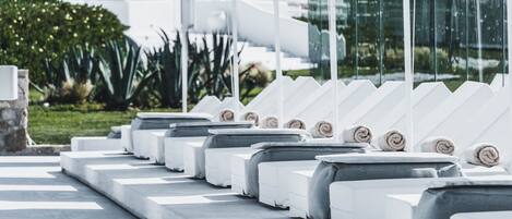 A row of white sunbeds with towels, a stone wall, and a glass facade reflecting the surroundings.